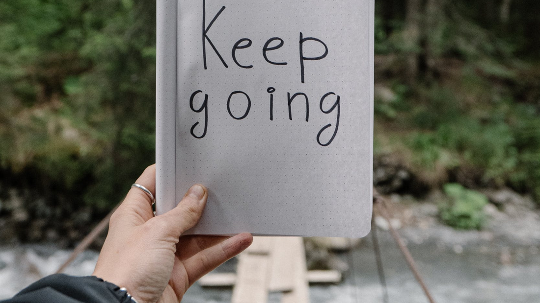 A person's hand holding open a notebook with 'Keep going' handwritten on a dotted page, with a natural, blurred background of a forest and a wooden footbridge.